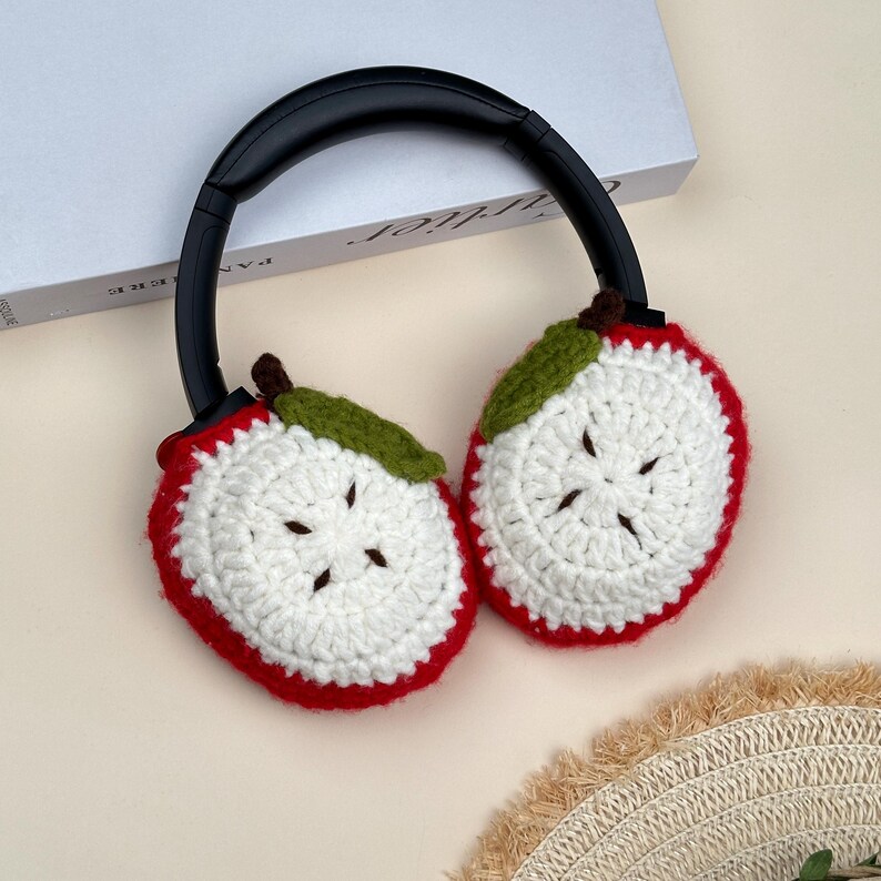 Puede incluir: Auriculares de crochet con cubiertas para los o&iacute;dos en forma de rodajas de manzana. Las cubiertas para los o&iacute;dos son blancas con bordes rojos, hojas verdes y tallos marrones. Los auriculares tienen una diadema negra.