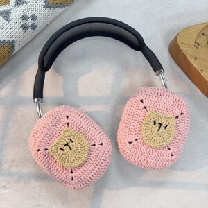 May include: Pink crocheted headphone covers with a brown bear face design. The covers are attached to a black headband with a mesh design.