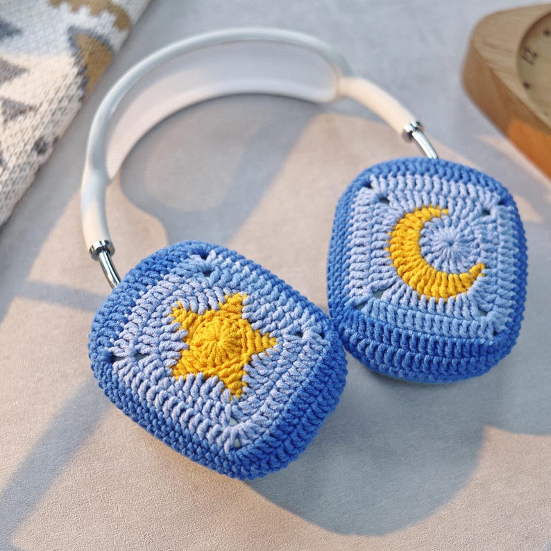 Puede incluir: Orejeras de crochet azul con una estrella amarilla en un lado y una luna creciente amarilla en el otro.