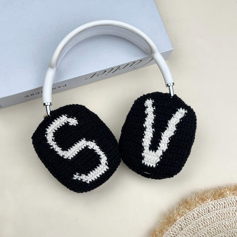 Puede incluir: Auriculares blancos y plateados con almohadillas negras. Las almohadillas est&aacute;n tejidas con las letras "S" y "V" en blanco. Los auriculares descansan sobre una superficie de color claro.