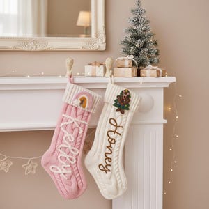 May include: Two cable-knit Christmas stockings, one pink with "Kelly" embroidered, and one cream with "Honey" embroidered. Each has a decorative top with a rainbow and a Christmas tree with a bear. The stockings hang from a white mantelpiece.