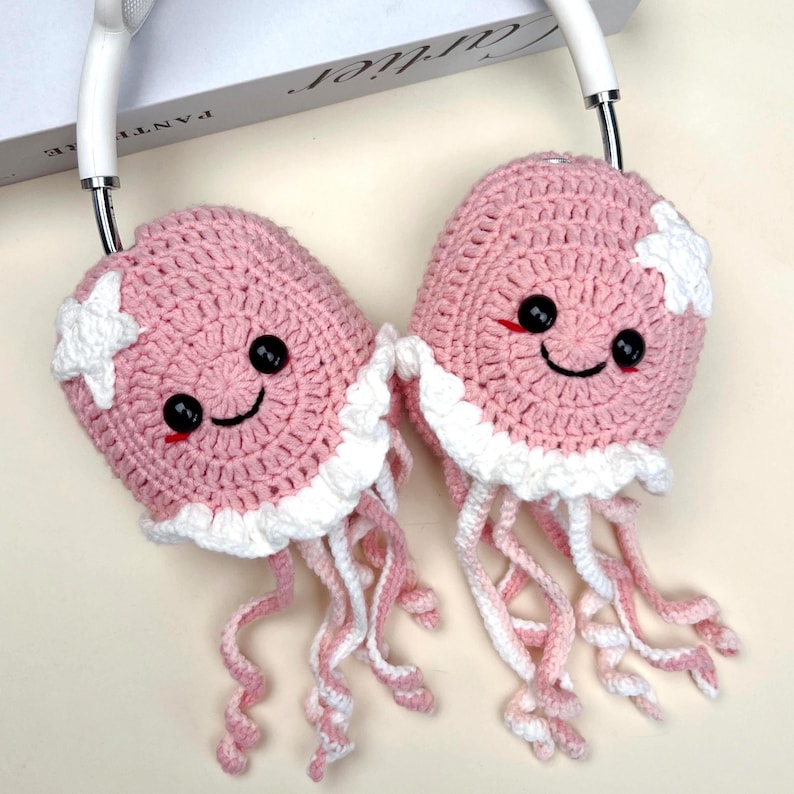 Puede incluir: Fundas de auriculares de ganchillo rosas y blancas con forma de medusas sonrientes. Cada funda tiene ojos de bot&oacute;n negros, una boca roja y un acento de estrella blanca. Las medusas tienen tent&aacute;culos blancos y rosas.