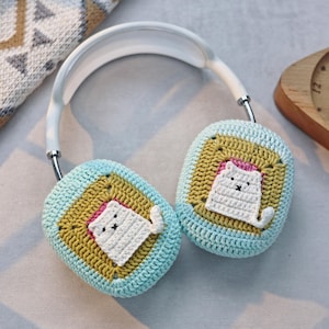 May include: White headphones with crocheted earmuffs featuring a cat design. The earmuffs are blue and yellow with a white cat in the center.