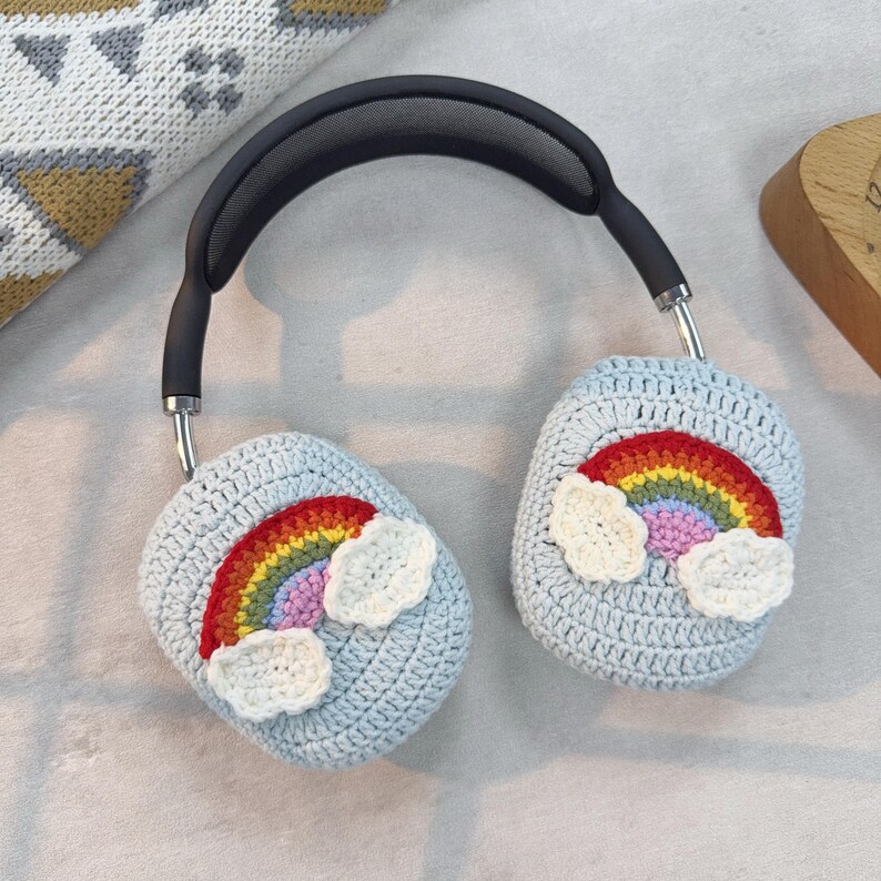 May include: Light blue crochet headphone covers with a rainbow and cloud design. The rainbow features red, orange, yellow, green, and purple colors. The headphones have a black headband and silver accents.