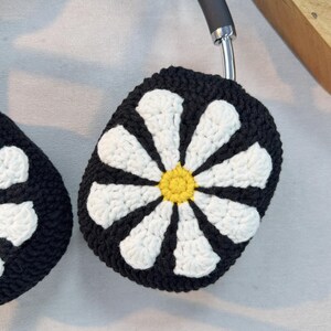K&ouml;nnte beinhalten: Schwarze und wei&szlig;e geh&auml;kelte Kopfh&ouml;rerbez&uuml;ge mit einem G&auml;nsebl&uuml;mchen-Design. Die G&auml;nsebl&uuml;mchen haben einen gelben Mittelpunkt.