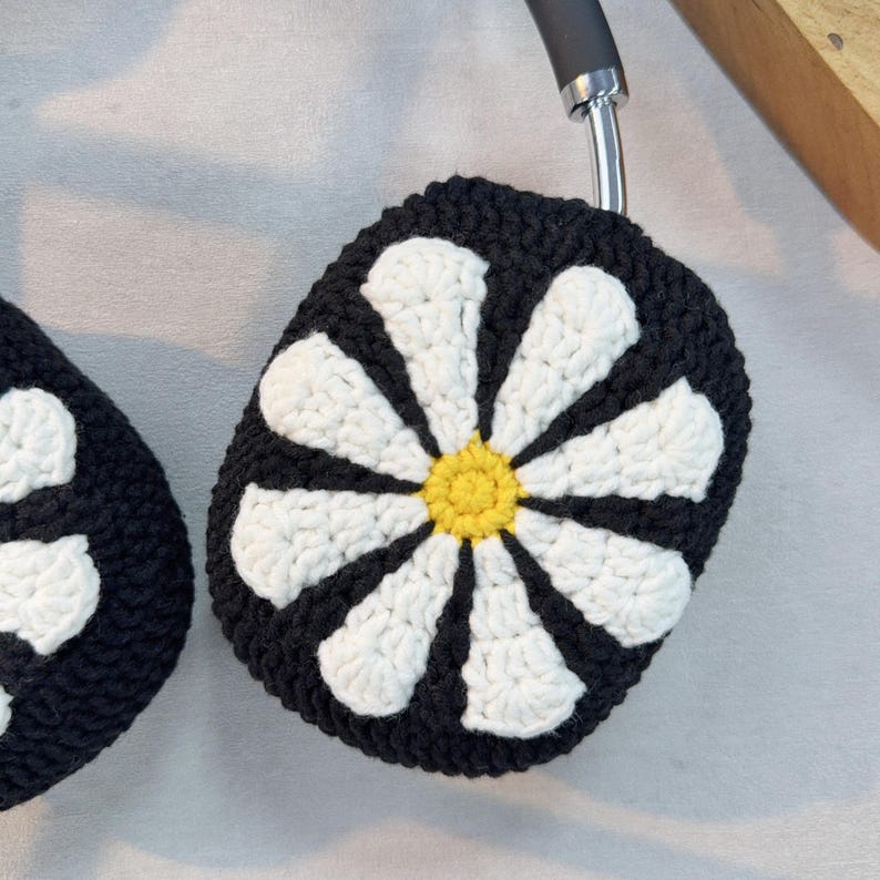 K&ouml;nnte beinhalten: Schwarze und wei&szlig;e geh&auml;kelte Kopfh&ouml;rerbez&uuml;ge mit einem G&auml;nsebl&uuml;mchen-Design. Die G&auml;nsebl&uuml;mchen haben einen gelben Mittelpunkt.