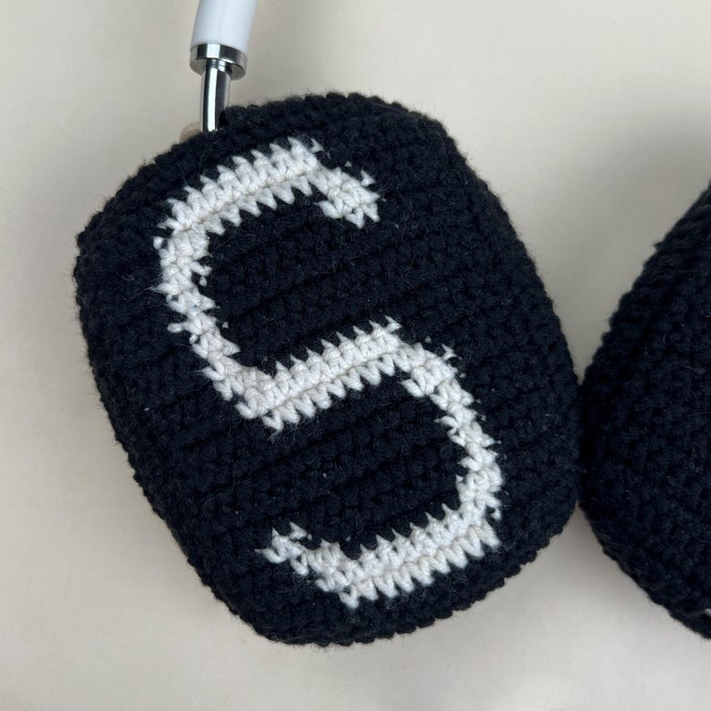 Puede incluir: Fundas de auriculares de ganchillo negras con un dise&ntilde;o de letra "S" blanca. Las fundas est&aacute;n hechas a mano y parecen estar hechas de hilo suave. Los auriculares tienen una diadema blanca.