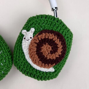 Pu&ograve; includere: Copri cuffie verdi all'uncinetto con un design a forma di lumaca. La lumaca ha un corpo bianco, un guscio marrone e beige e sorride. Le cuffie sono bianche.