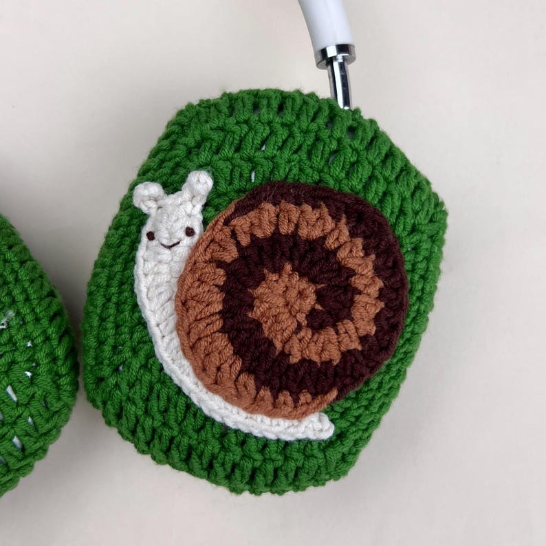 Pu&ograve; includere: Copri cuffie verdi all'uncinetto con un design a forma di lumaca. La lumaca ha un corpo bianco, un guscio marrone e beige e sorride. Le cuffie sono bianche.