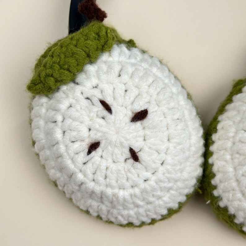 Puede incluir: Primer plano de una rodaja de manzana de ganchillo hecha a mano. La rodaja de manzana es blanca con detalles de semillas marrones y una hoja y tallo de ganchillo verdes. Los bordes est&aacute;n delineados con hilo verde.