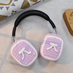 May include: Purple crocheted earmuffs with white bows on a light gray surface.
