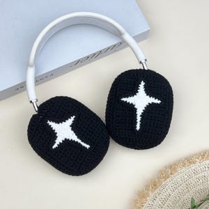 May include: Black crochet headphones with a white starburst design on each ear cup. The headband is white, and the headphones rest on a light-colored surface. The ear cups are oval-shaped.
