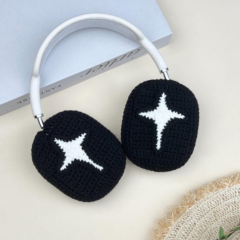 May include: Black crochet headphones with a white starburst design on each ear cup. The headband is white, and the headphones rest on a light-colored surface. The ear cups are oval-shaped.