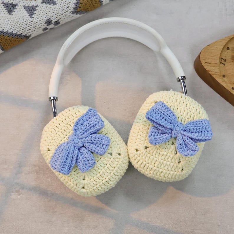 May include: White headphones with crocheted earmuffs. The earmuffs are yellow with a blue crocheted bow on each side.