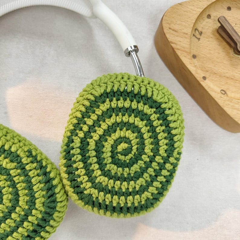 Puede incluir: Orejeras de crochet verde y verde claro con un patr&oacute;n circular. Las orejeras est&aacute;n unidas a una diadema blanca.