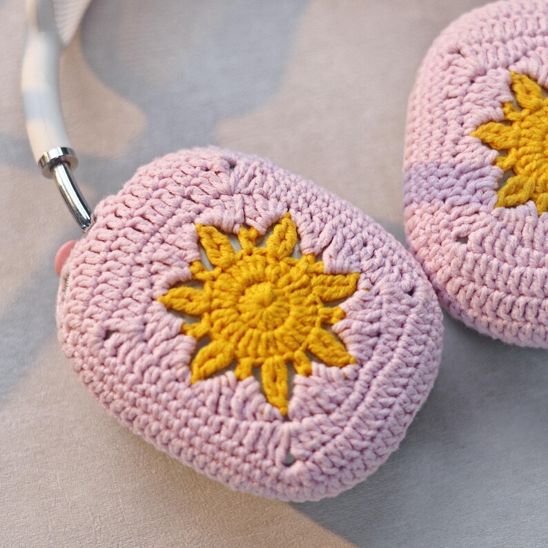 K&ouml;nnte beinhalten: Ein Paar rosa H&auml;kel-Kopfh&ouml;rerbez&uuml;ge mit gelben H&auml;kelblumen-Designs.