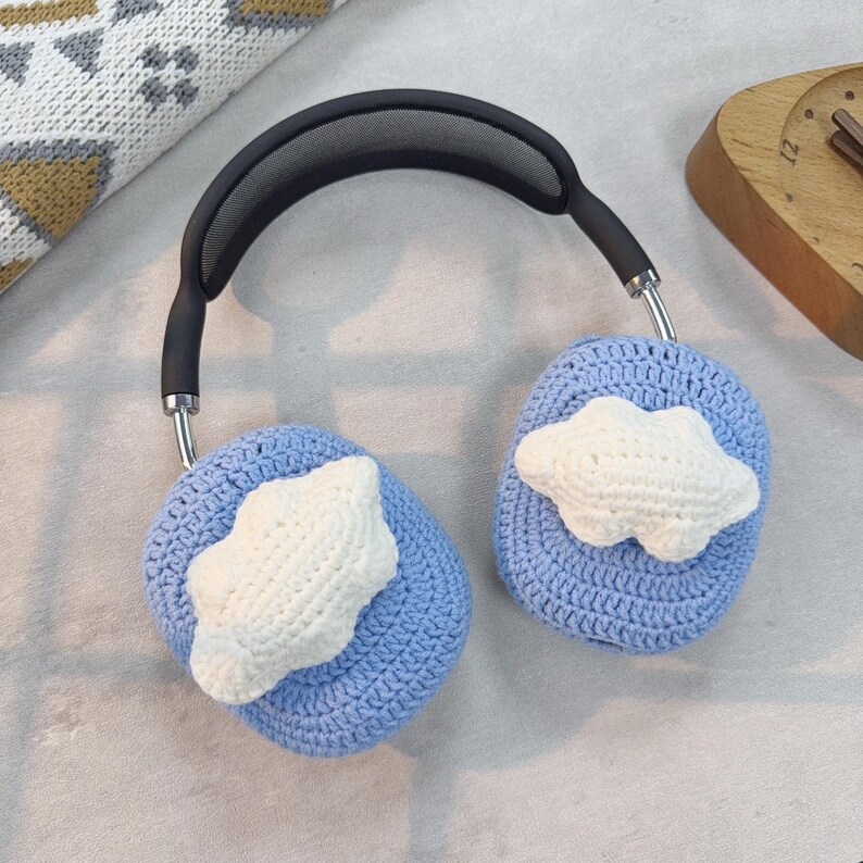 May include: Blue crocheted headphone covers with white cloud designs. The covers are attached to a black headband with a silver metal frame.