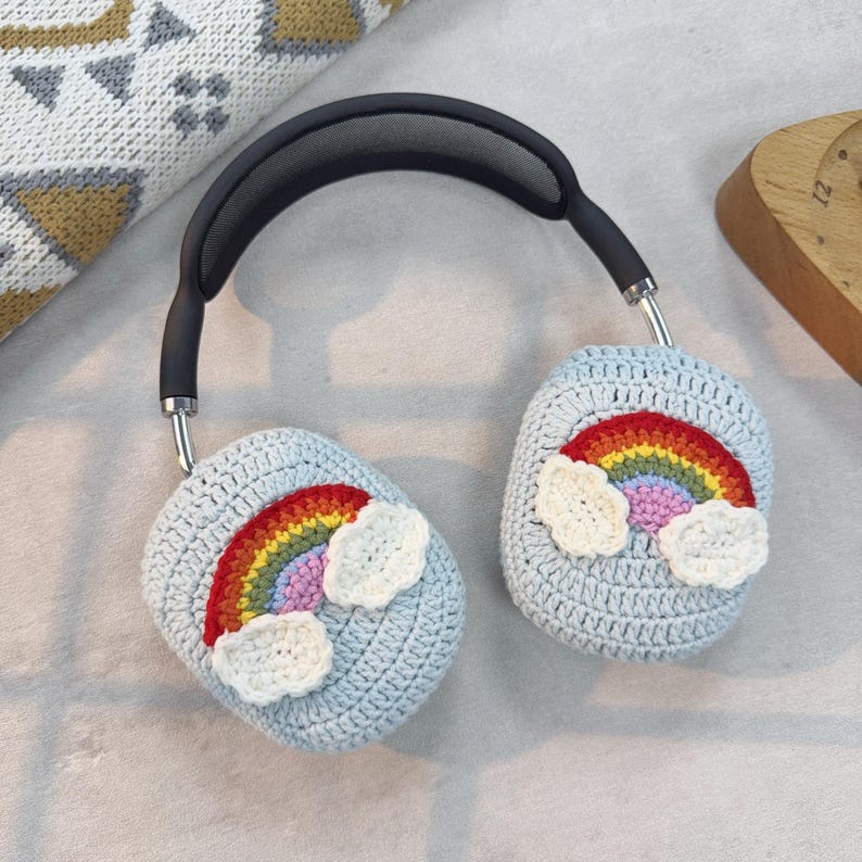 May include: Over-ear headphones with light blue crochet ear covers. Each cover features a colorful rainbow and white cloud design. The headphones have a black headband and silver accents.
