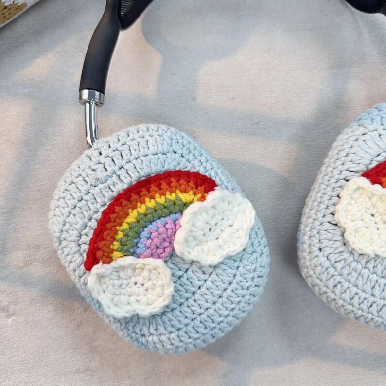 May include: Crocheted light blue headphone covers with a rainbow and cloud design. The rainbow features red, orange, yellow, green, blue, and purple colors. The clouds are white. The headphones are black and silver.