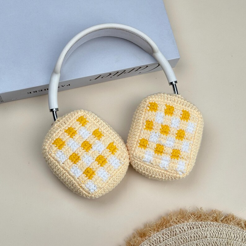May include: White and silver headphones with cream-colored, crocheted ear covers. The ear covers feature a yellow and white checkered pattern. The headphones are resting on a book.