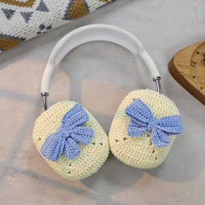 May include: A pair of white headphones with crocheted earmuffs. The earmuffs are light yellow with a blue crocheted bow on each one.