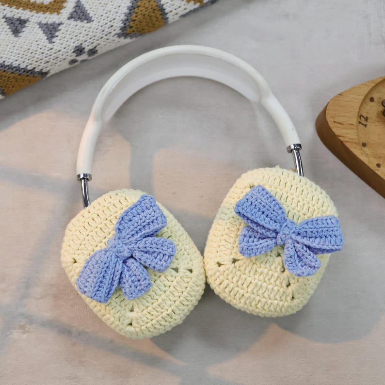 May include: A pair of white headphones with crocheted earmuffs. The earmuffs are light yellow with a blue crocheted bow on each one.