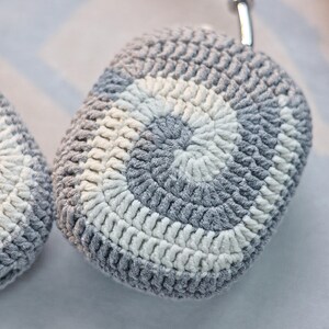 May include: Grey and white crocheted case for wireless headphones. The case has a circular pattern with a white centre and grey outer ring.