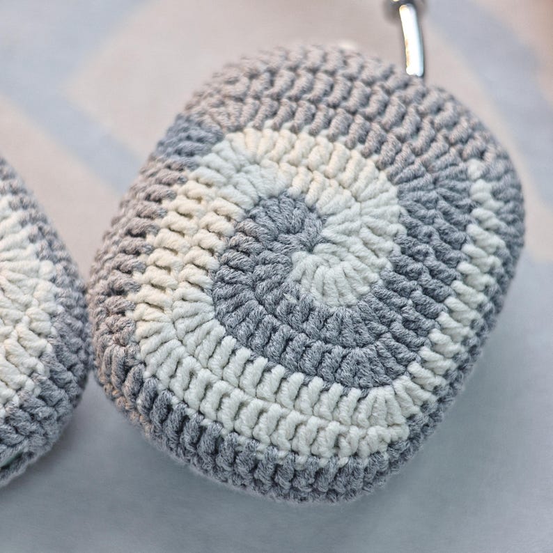 May include: Grey and white crocheted case for wireless headphones. The case has a circular pattern with a white centre and grey outer ring.