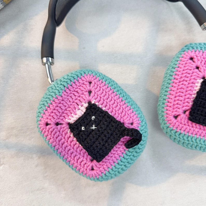 May include: A pair of pink and teal crocheted headphone covers with a black crocheted cat design.