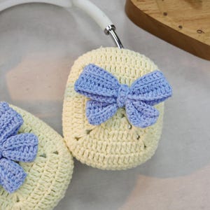 May include: A pair of crocheted headphone covers with a light yellow background and a blue bow on each cover.