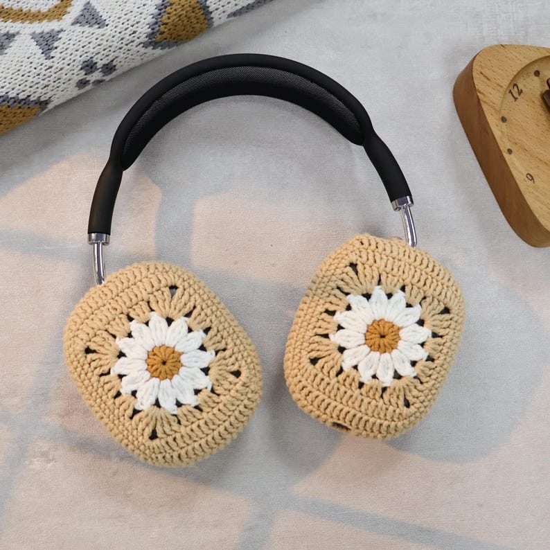 Puede incluir: Fundas para auriculares de crochet beige con un dise&ntilde;o de margarita blanca. Las fundas est&aacute;n unidas a una diadema negra.