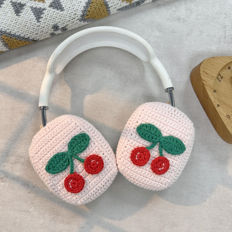 May include: White headphones with pink crocheted earmuffs featuring embroidered cherries. The earmuffs have a light pink background with green leaves and red cherries.