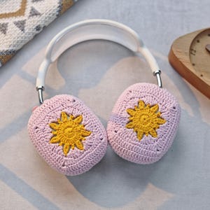 K&ouml;nnte beinhalten: Ein wei&szlig;er Kopfh&ouml;rer mit geh&auml;kelten Ohrpolstern. Die Ohrpolster sind rosa mit einem gelben geh&auml;kelten Blumendesign.