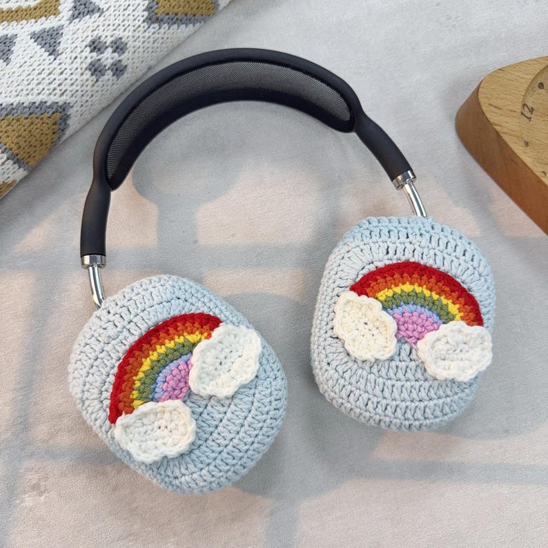 May include: Headphones with light blue crochet ear covers. Each cover features a colorful rainbow design with white cloud accents. The headphones have a black headband and silver metal arms.