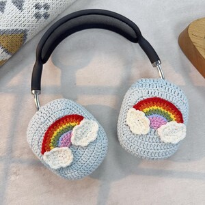 May include: Headphones with light blue crochet ear covers, each adorned with a colorful rainbow and white cloud design. The headphones have a black headband and silver accents. The ear covers are a handmade, whimsical accessory.