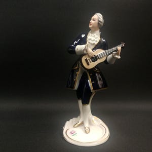 Può includere: Statuina in porcellana di un uomo in abiti del XVIII secolo, che suona un liuto. La figura indossa un cappotto blu navy con finiture dorate, una camicia bianca con volant e una parrucca bianca. La base è bianca con una rosa rosa.