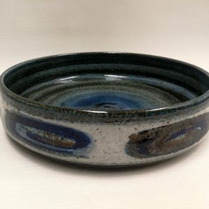 May include: A handmade ceramic bowl with a wide, shallow design. The interior features concentric circles of blue and teal, while the rim is a darker blue. The exterior has a white base with blue and brown abstract designs.