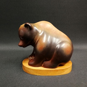 Könnte beinhalten: Eine Keramik-Bärenfigur in Braun- und Beigetönen, die auf einem runden Holzsockel sitzt. Der Bär hat eine glänzende Oberfläche, und der Holzsockel ist hellbraun. Die Figur ist ein dekorativer Gegenstand.