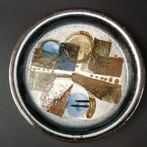 Puede incluir: Un plato redondo de cerámica con interior blanco y borde marrón oscuro. El plato presenta un diseño abstracto con formas marrones, azules y beige. El diseño del plato incluye una serie de puntos y líneas. Es un artículo decorativo.