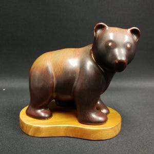 Pode incluir: Estatueta de urso de cerâmica castanha com acabamento brilhante, sobre uma base de madeira. O urso tem cabeça e corpo castanho escuro, com detalhes castanhos claros. Os olhos são pequenos e dourados. É um objeto decorativo.