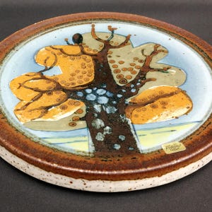 Op de afbeelding: Een rond keramisch bord met een gestileerd boomontwerp met een donkerbruine stam en takken, geel en beige gebladerte met cirkelvormige patronen en een lichtblauwe achtergrond. De rand is donkerbruin met een gespikkelde witte onderkant.