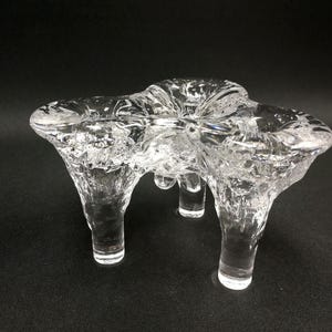 Puede incluir: Un candelabro de vidrio transparente con tres portavelas y tres patas. El candelabro tiene un diseño texturizado y orgánico. El candelabro está sobre una superficie negra.