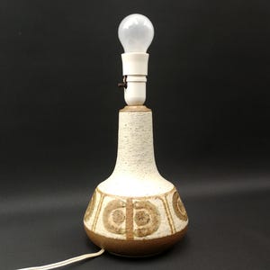 May include: A ceramic table lamp with a bulb. The lamp has a cream-colored, textured body with brown circular patterns and a brown base. The lamp is plugged in and ready to use.
