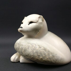 May include: A ceramic sculpture of a white arctic fox with gray markings, sitting with its head turned to the side.