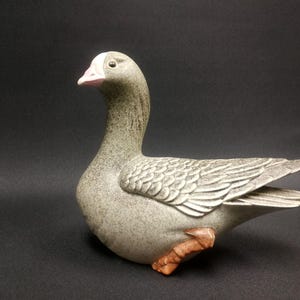 May include: A ceramic goose figurine with a gray and white speckled finish. The goose has a pink beak and orange feet.