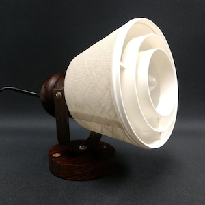 May include: A wall-mounted lamp featuring a dark brown wooden circular base and bracket. The lamp head consists of a conical shade with a textured off-white fabric exterior and a series of concentric white plastic rings inside, with a black cord.