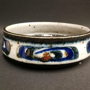 May include: A round ceramic bowl with a low profile, featuring a white base glaze speckled with brown dots. The exterior is decorated with a horizontal band of abstract blue and dark green swirls, finished with a dark brown rim and interior edge.