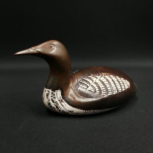 May include: A wooden sculpture of a loon bird. The bird has a brown body with white and black detailing on the underside and wings. The loon has a long, pointed beak and is set against a black background.