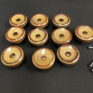 Puede incluir: Un juego de nueve pomos redondos de cerámica para armarios con un acabado brillante en color tostado y un acento circular de color marrón rojizo oscuro alrededor del centro. Cada pomo tiene un orificio central. Incluye varios tornillos de metal.