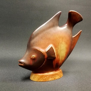 Könnte beinhalten: Eine Keramik-Fischfigur mit brauner und orangefarbener Glasur. Der Fisch steht auf einem Holzsockel.